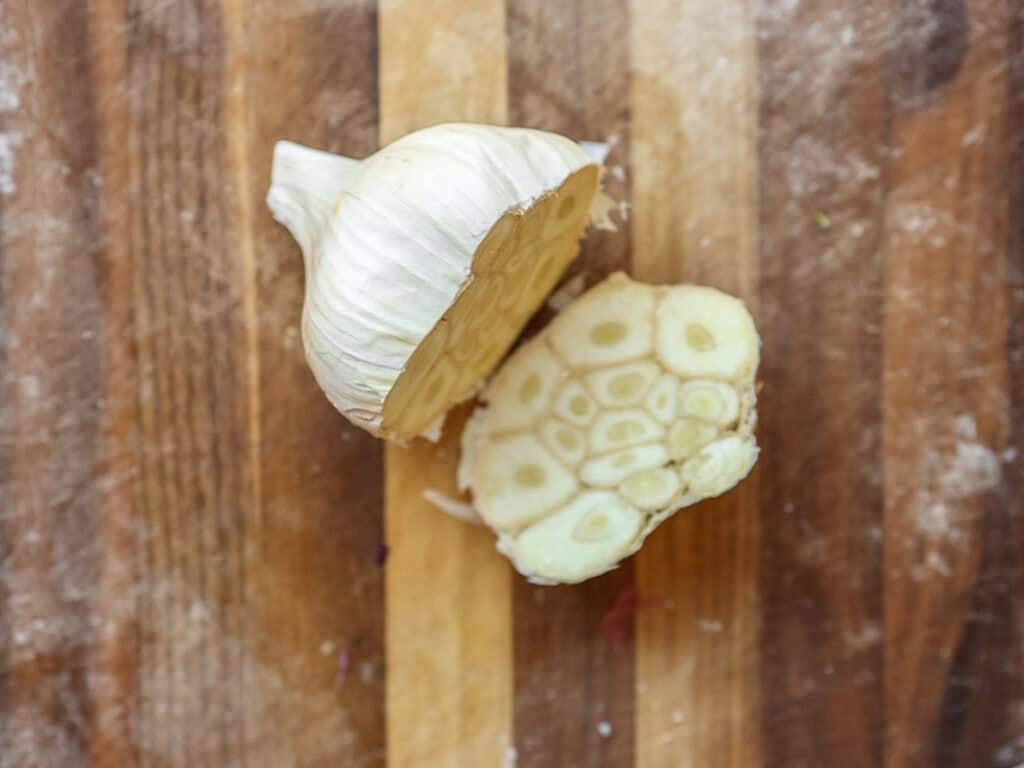 A halved garlic bulb on a wooden cutting board, showing the cross-section with multiple cloves. The garlics layers and cloves are visible, surrounded by a dusting of flour on the board.